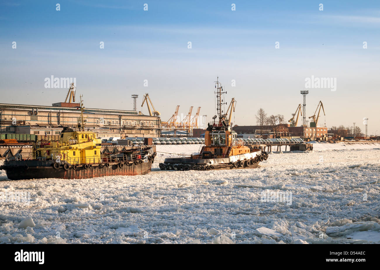 Small tug boat goes with vessel through icy channel in harbor of St.Petersburg cargo port Stock Photo