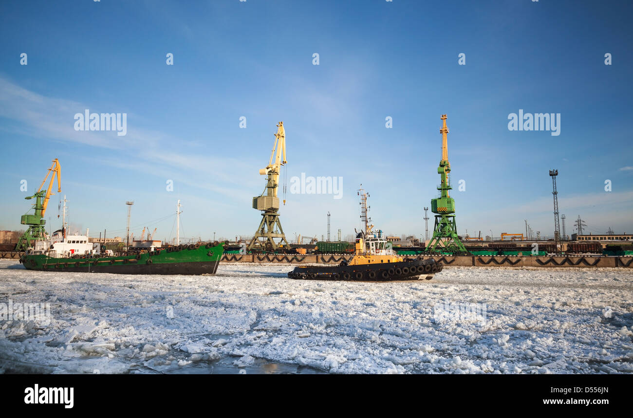 Small tug boat goes with vessel through icy channel in harbor of St.Petersburg cargo port Stock Photo