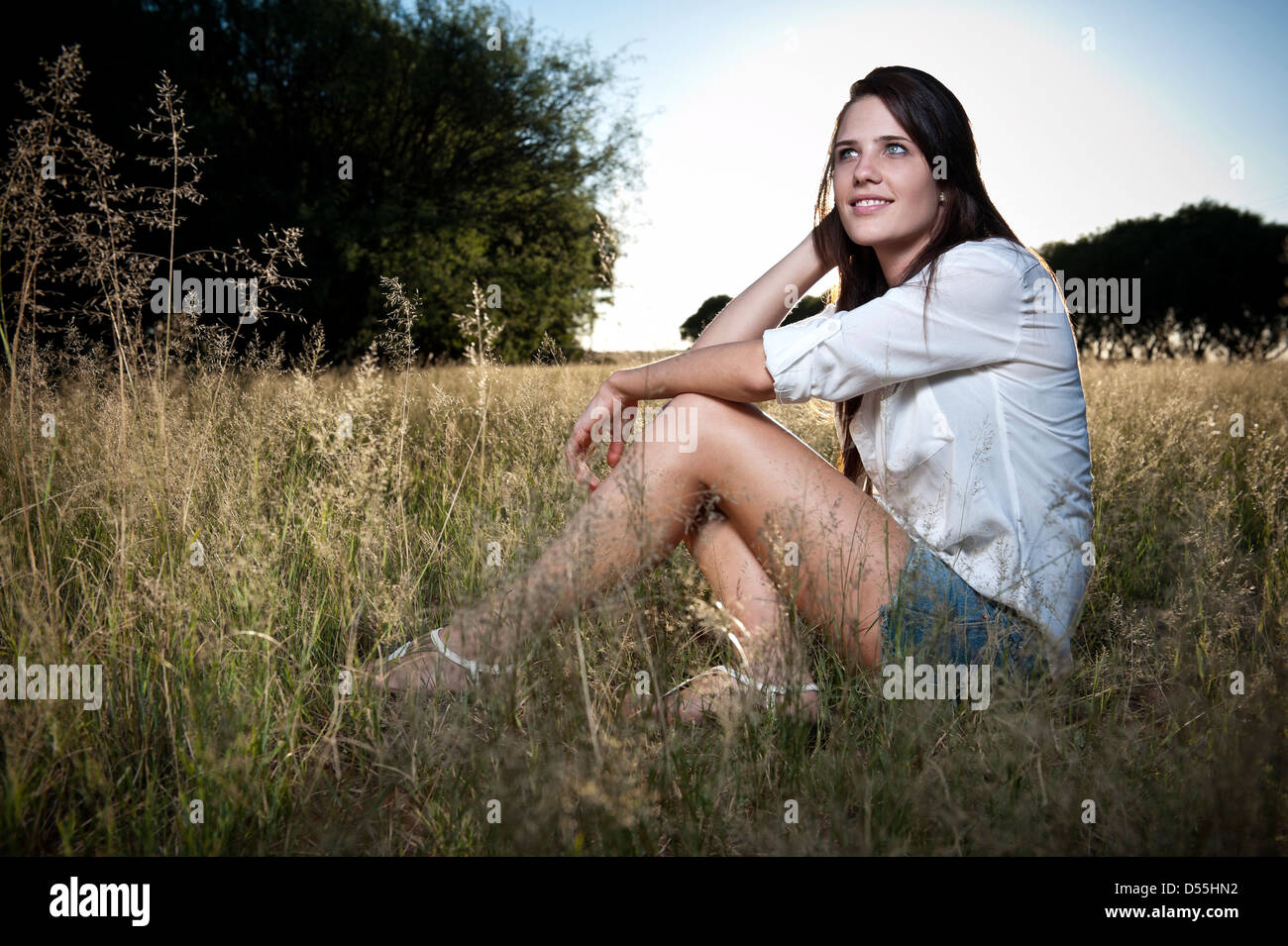 Gorgeous, beautiful, pretty, Caucasian brunette, with blue eyes, sitting in a field with tall grass at sunset with white shirt. Stock Photo