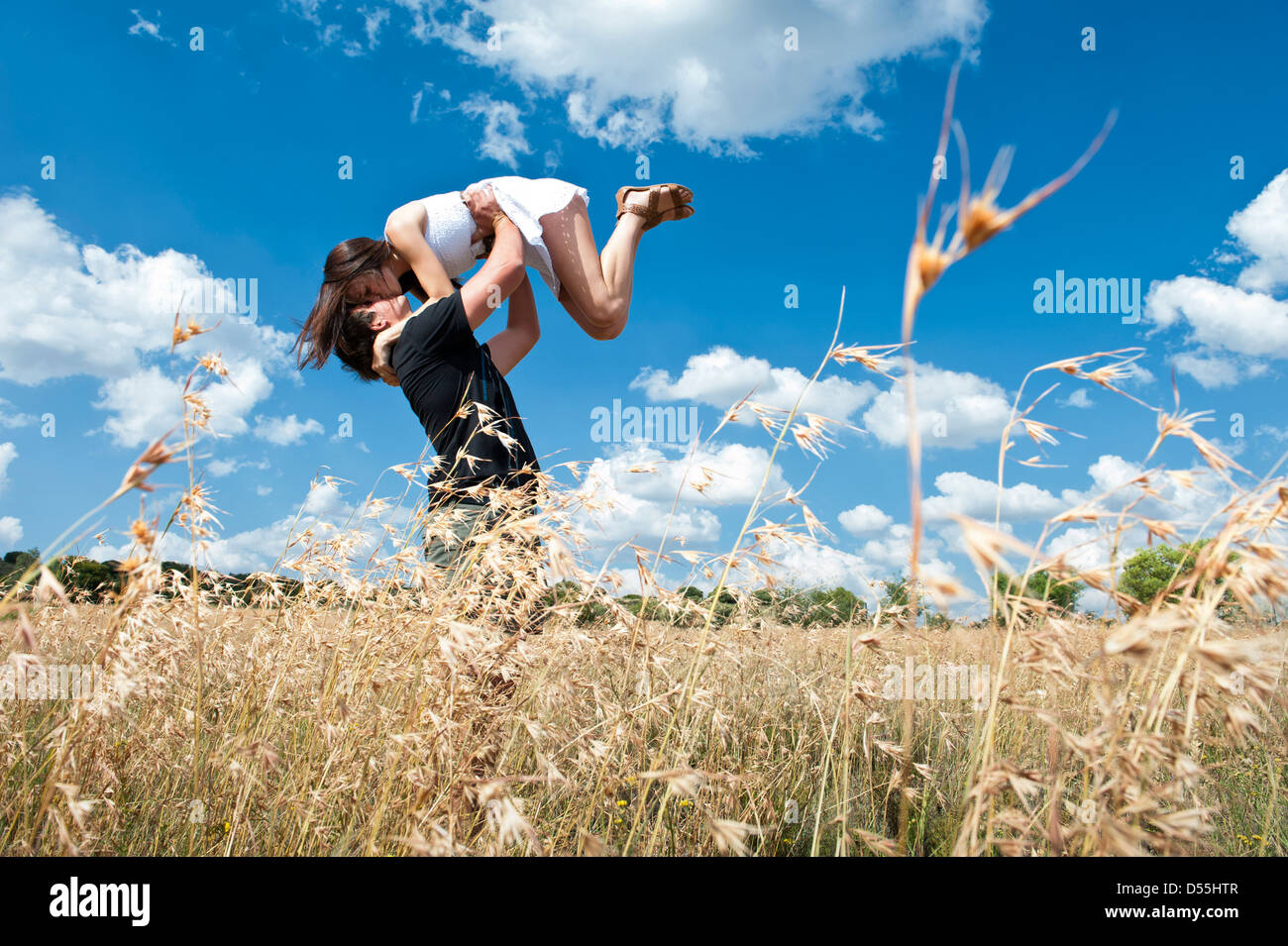 Happy young couple standing in open field of long grass with the young man lifting the gorgeous girl into the air, kissing. Stock Photo