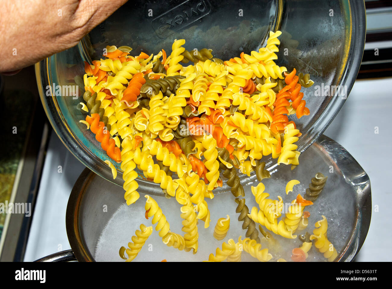 Pouring bowl dry uncooked tricolour rotini pasta into pot rapidly boiling water Stock Photo
