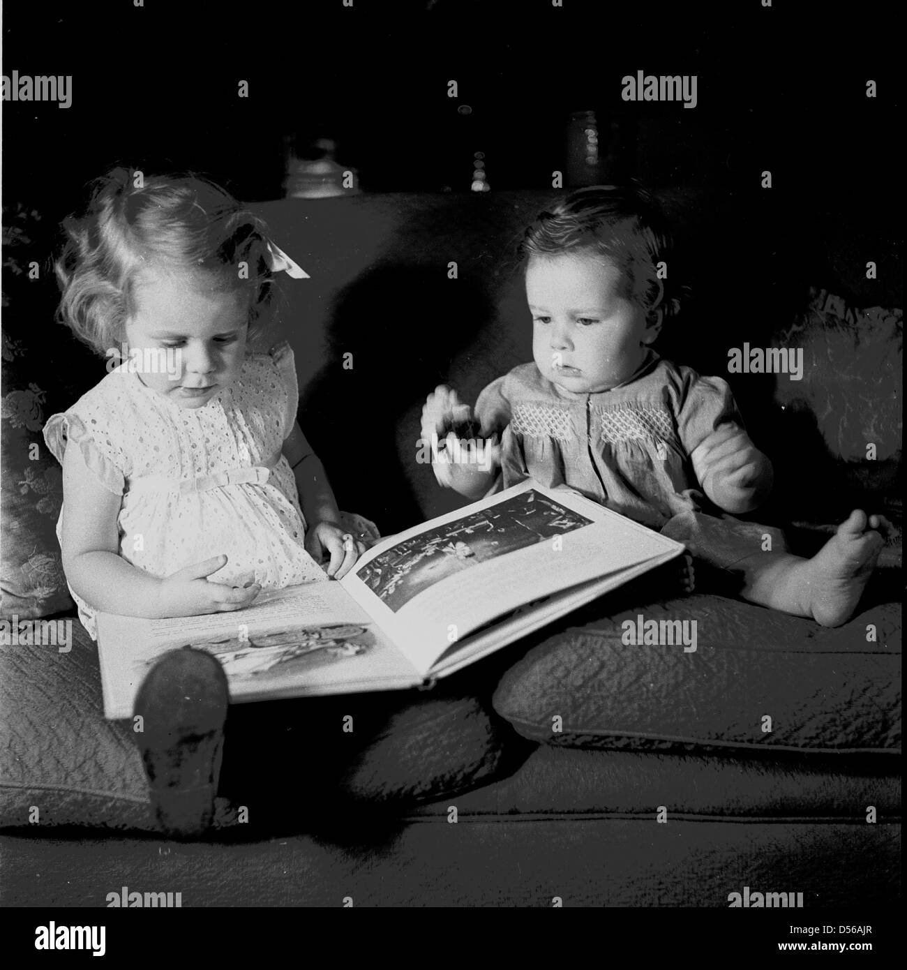 Historical 1950s. England. Little girl looks at a book while sitting on sofa next to baby girl. Stock Photo