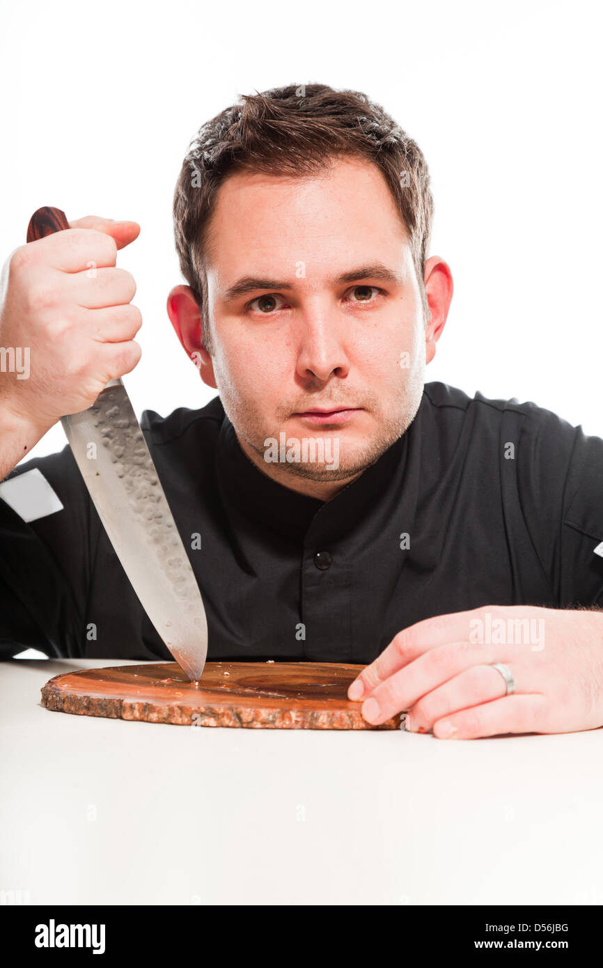 Young male cook with black jacket knife and wooden plate against white ...