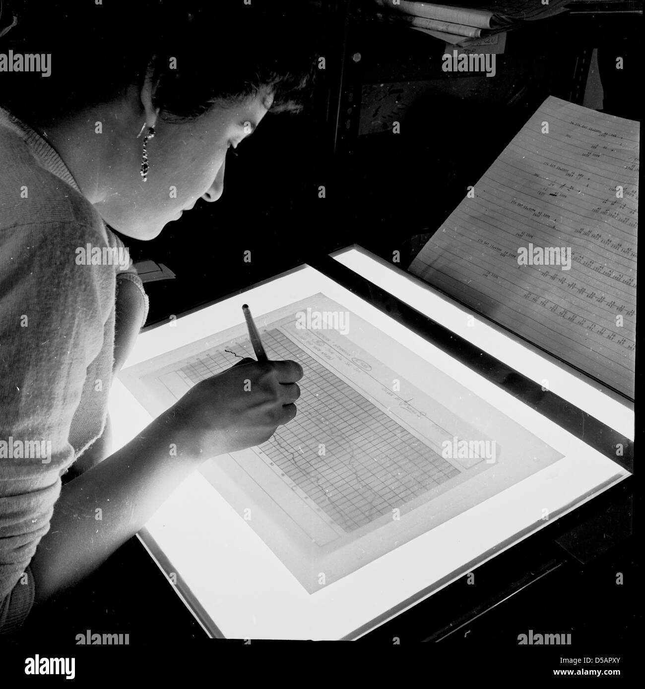 Historical picture, 1950s of a working woman using a lightbox to analyse figures on a graph against another graph. Stock Photo