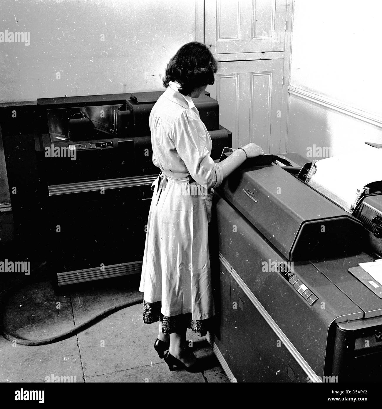 Historical picture from late 1950s of a woman working in an office using a piece of accounting equipment of the era, a large data processor and printing machine. Known as 'tabulators', they would read decks of cards and print selected portions of their contents, also adding up columns of numbers and printing totals.  Complex and cumbersome machines, they are considered the forerunners to early computers. Stock Photo
