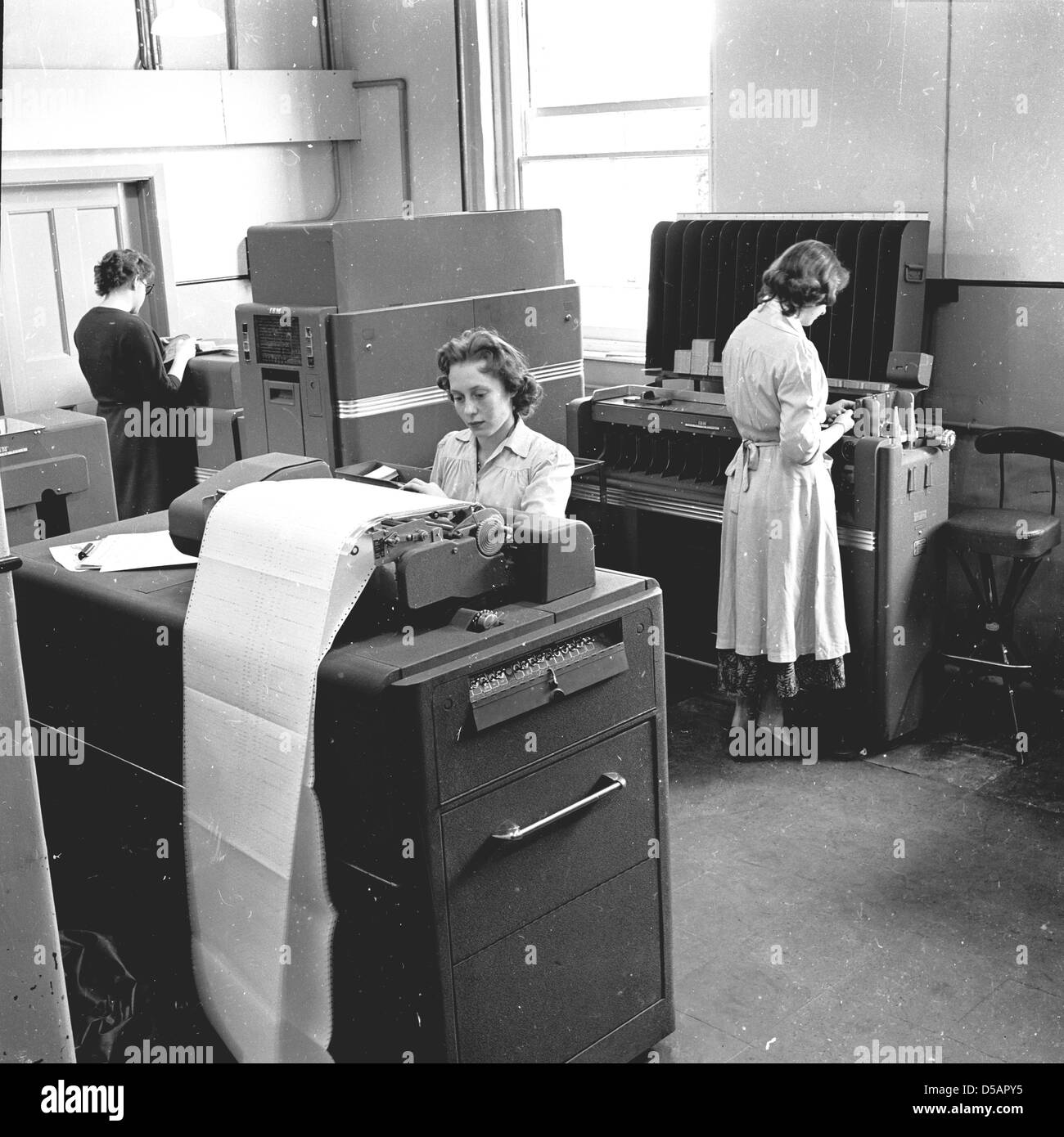 Historical picture from the late 1950s of three young women working in a company office using the large and cumbersome data processing machines and electronic printers of the era. One lady is working at a tabulator, an early data processing machine, used for processing data held on punched cards, while another lady is at what was referred to as a collator, a machine that sorted the punched cards. These machines were the forerunners of the early computers. Stock Photo