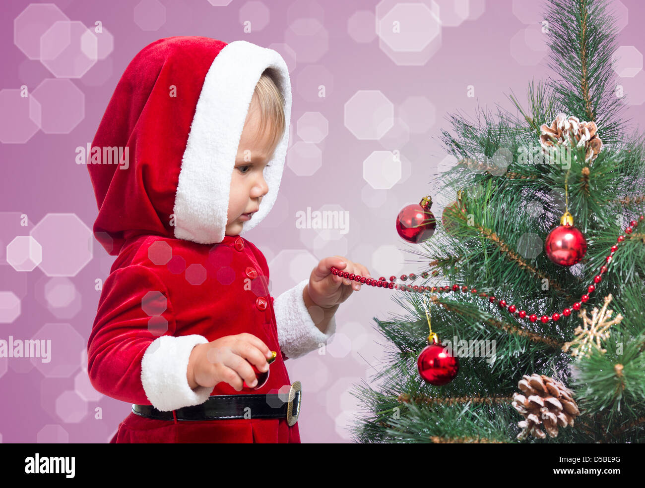 pretty Santa Claus baby decorating Christmas tree on bright festive background Stock Photo