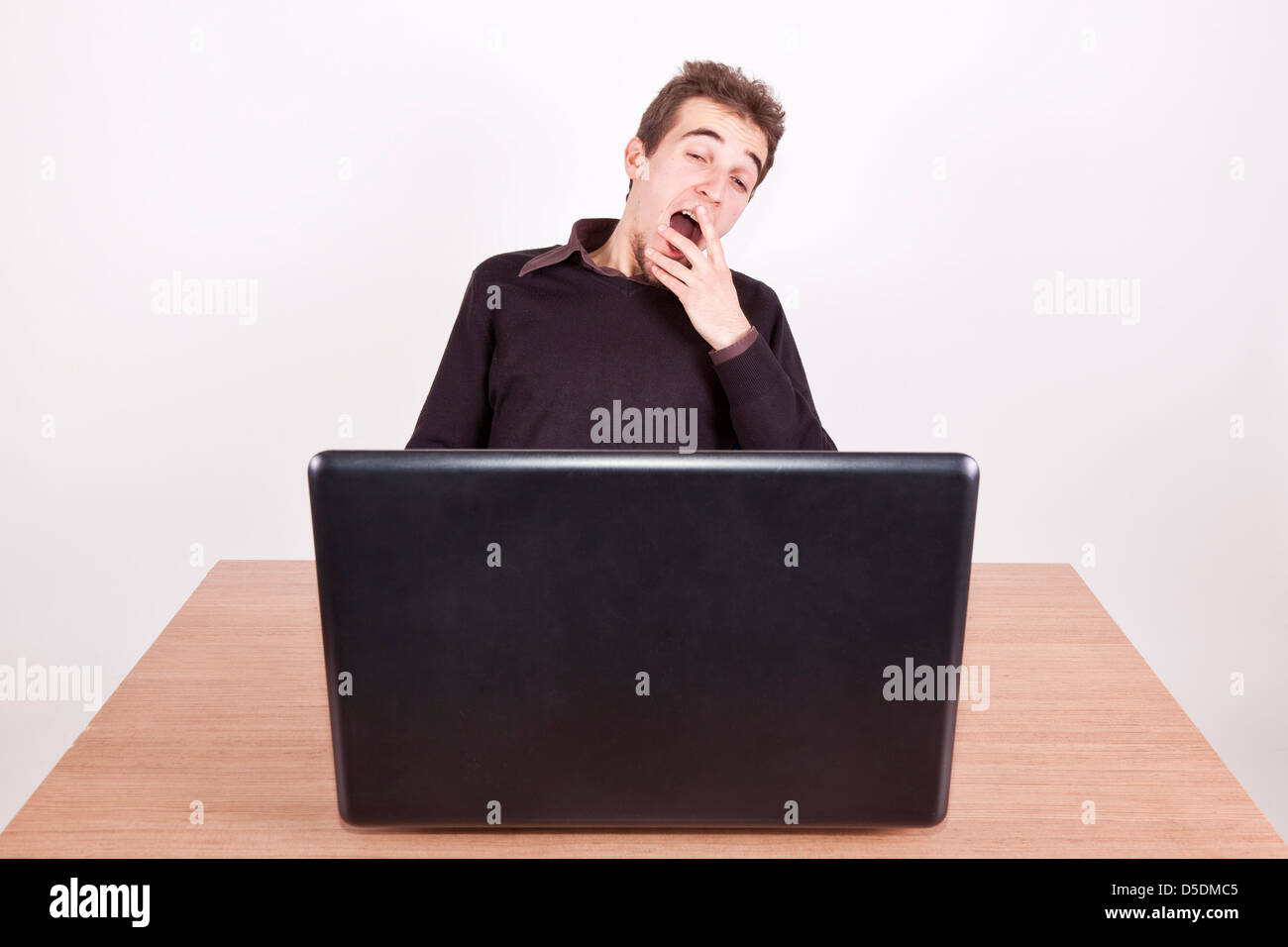 sleepy young man yawning at a desk with a laptop in front of him Stock Photo