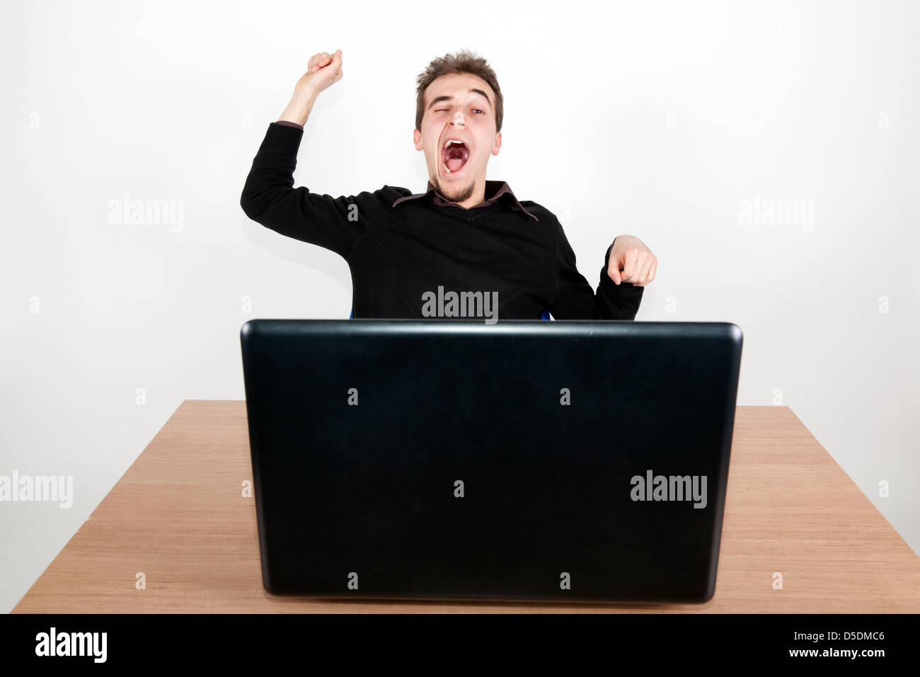 young man yawning at a desk with a laptop in front of him Stock Photo