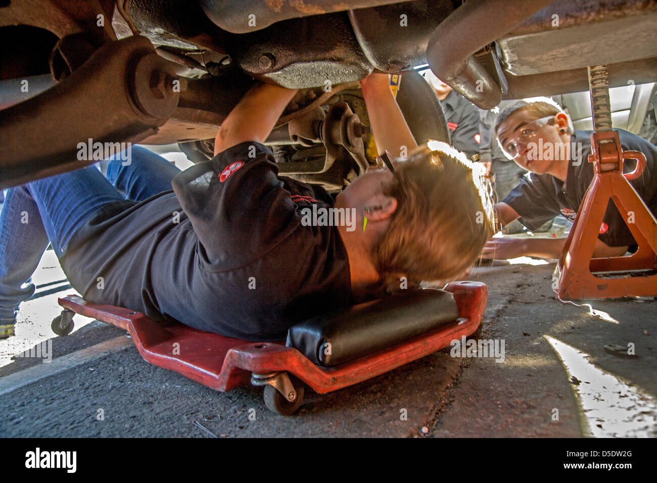 Wearing safety glasses a Caucasian teenage girl and boy work underneath a car engine in auto shop class in San Clemente, CA. Stock Photo