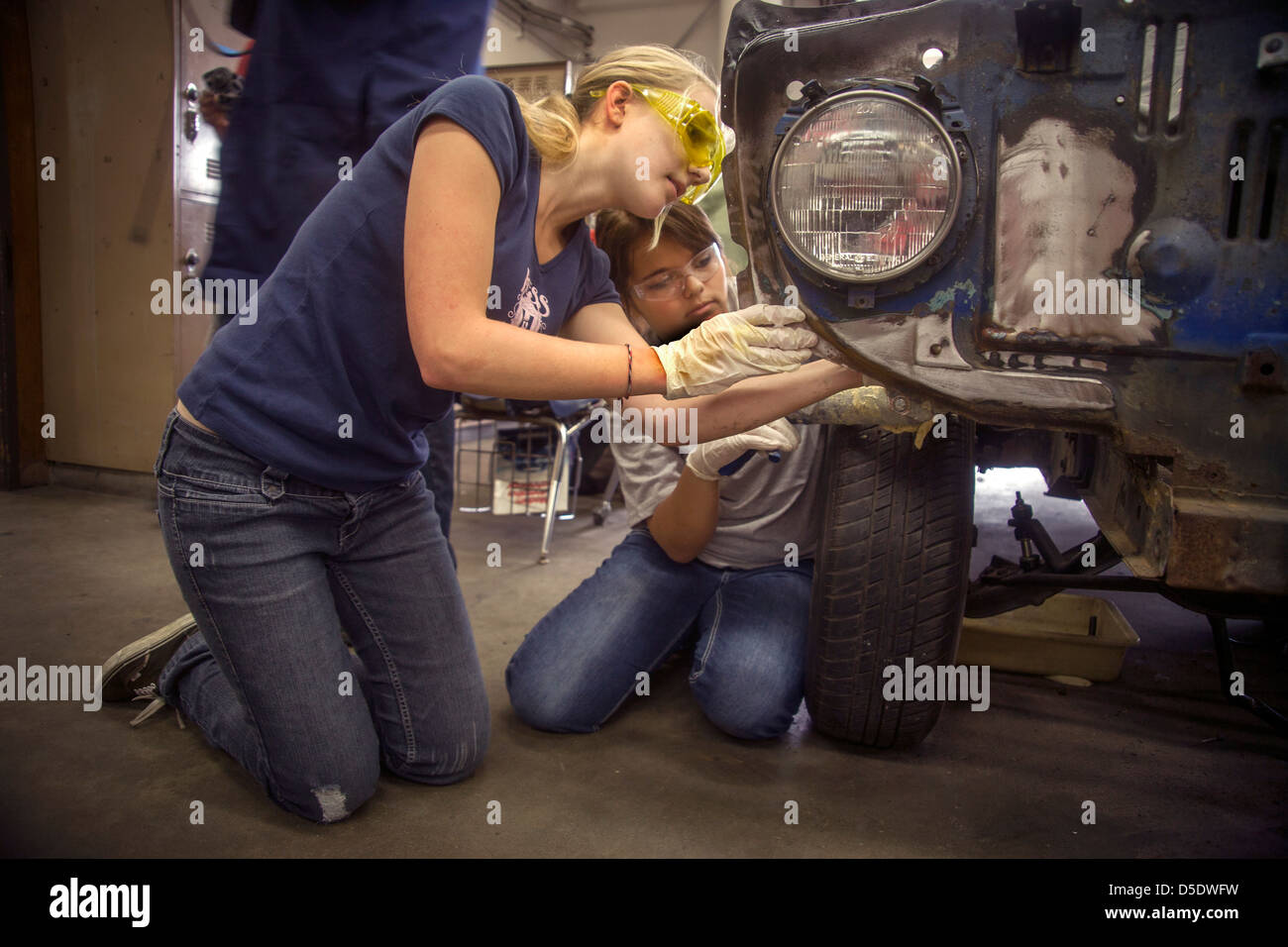 Wearing safety glasses two teenage girls work together on a car in auto