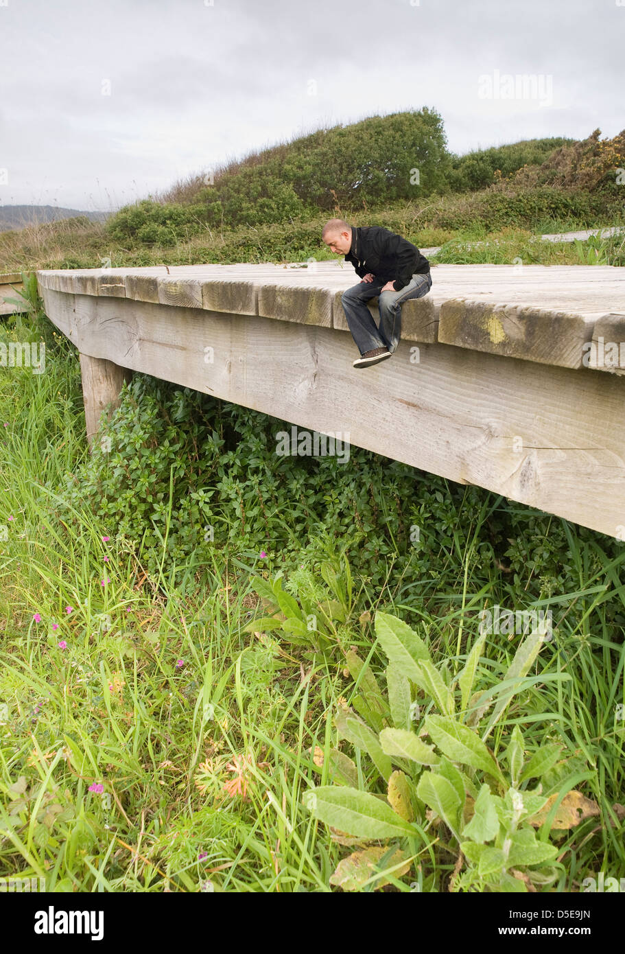 Photomontage of a young man sitting on a wooden walkway in nature and looking down. Man is small compared to the surrounding env Stock Photo