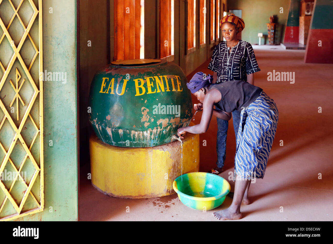 two woman drink holy water from a barrel upon the entrance door of the catholic cathedral in Bobo-Dioulasso, Burkina Faso Stock Photo