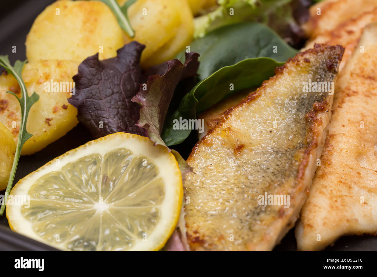 Fried perch filets with potatoes, lemon and salad in a black plate Stock Photo