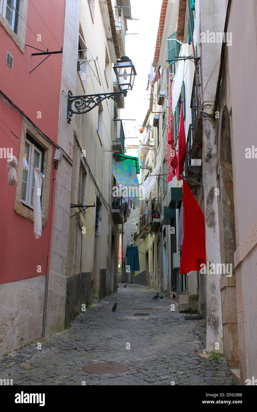 Narrow cobblestone alley street in Alfama, Lisbon, Portugal, with colorful laundry hanging from windows above Stock Photo