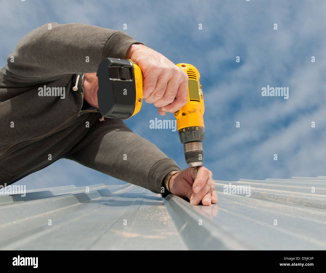 Man using an electric drill to fasten down a metal roof, view from below, up against party cloudy sky Stock Photo