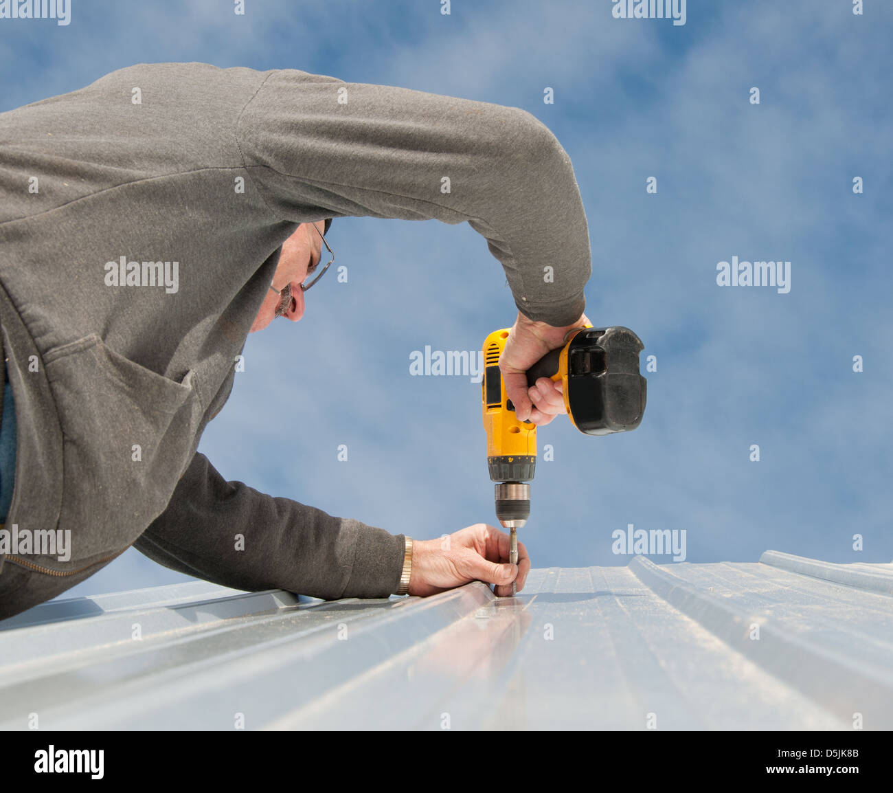 Man fastening down a metal roof with screws; with cloudy sky background Stock Photo