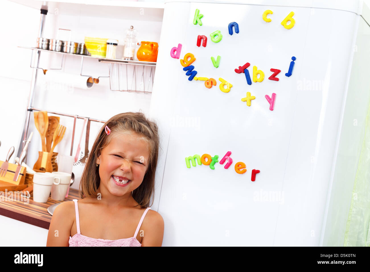 Little girl making funny face in front of the fridge with color letter magnets on it Stock Photo