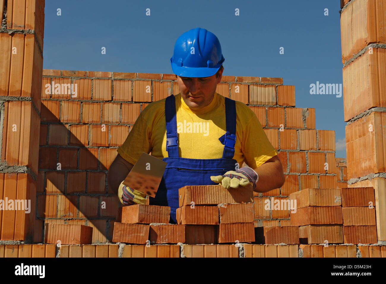 Mason in blue helmet and coveralls laying bricks of house wall Stock Photo