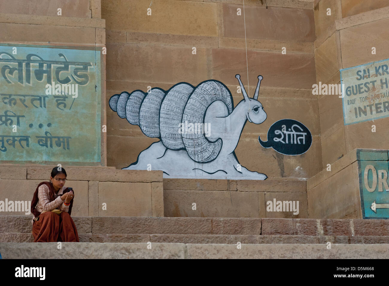 A woman using a mobile phone, sits under graffiti of a snail. Varanasi, India. Stock Photo