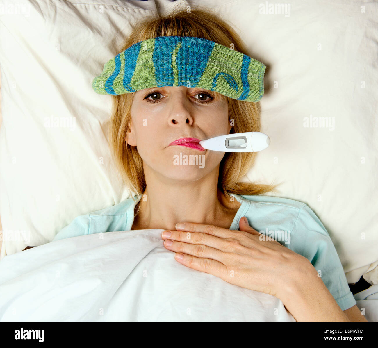 Woman sick in bed or hospital with thermometer in mouth and washcloth on face Stock Photo