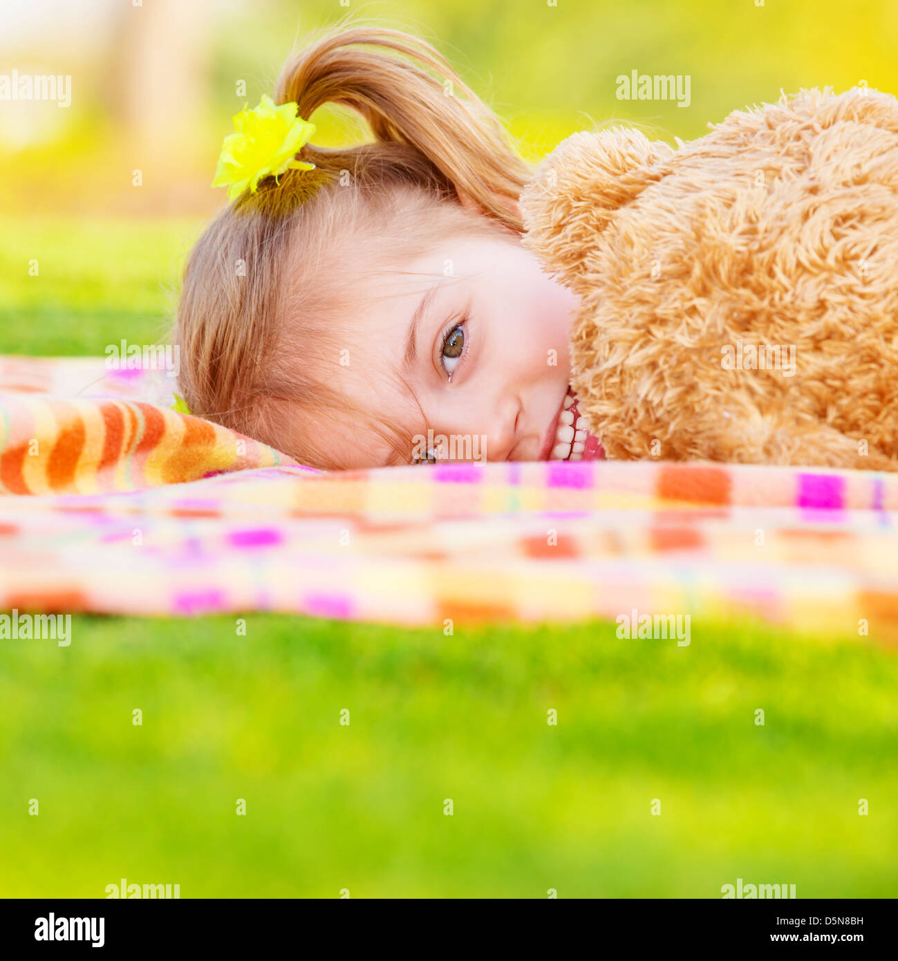 Pretty small girl lying down on green field, playing game with big brown teddy bear, cute child enjoying spring nature Stock Photo