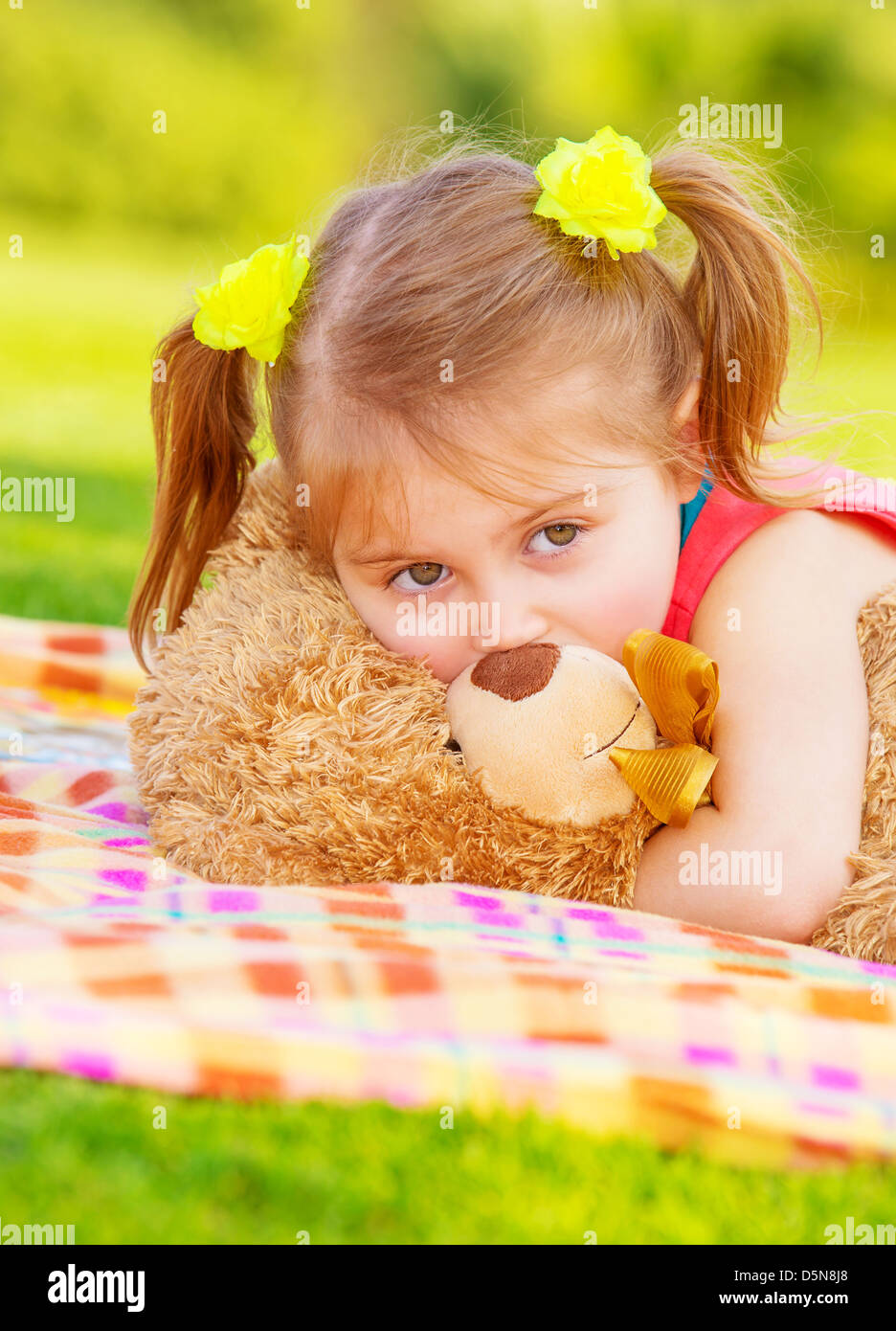 Closeup portrait of beautiful little girl having fun outdoors in spring time, lying down on green grass, hugging soft toy teddy Stock Photo