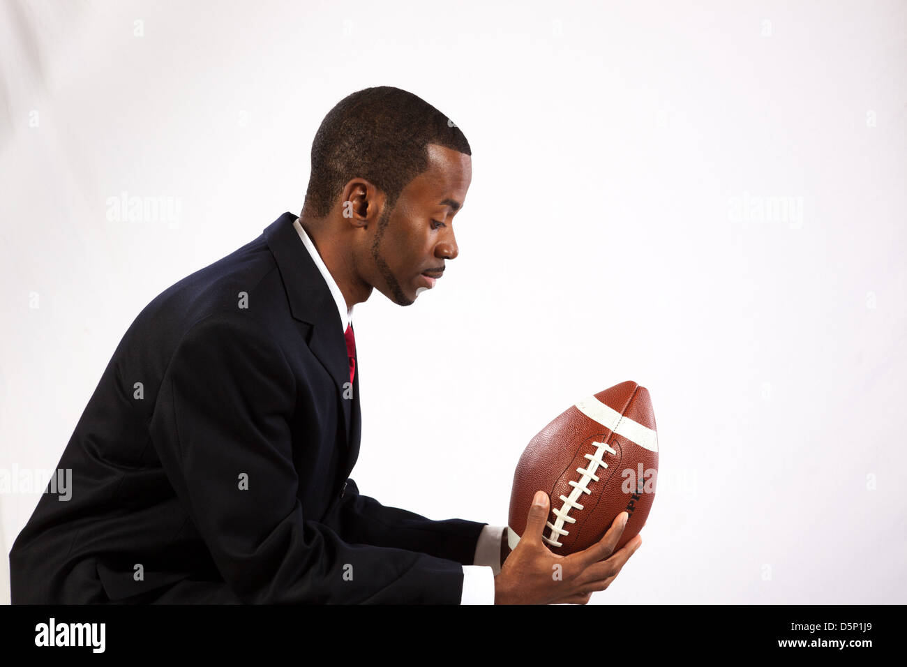 Handsome black man wearing a dark suit, white shirt and red tie, holding an American football and remembering his youth Stock Photo
