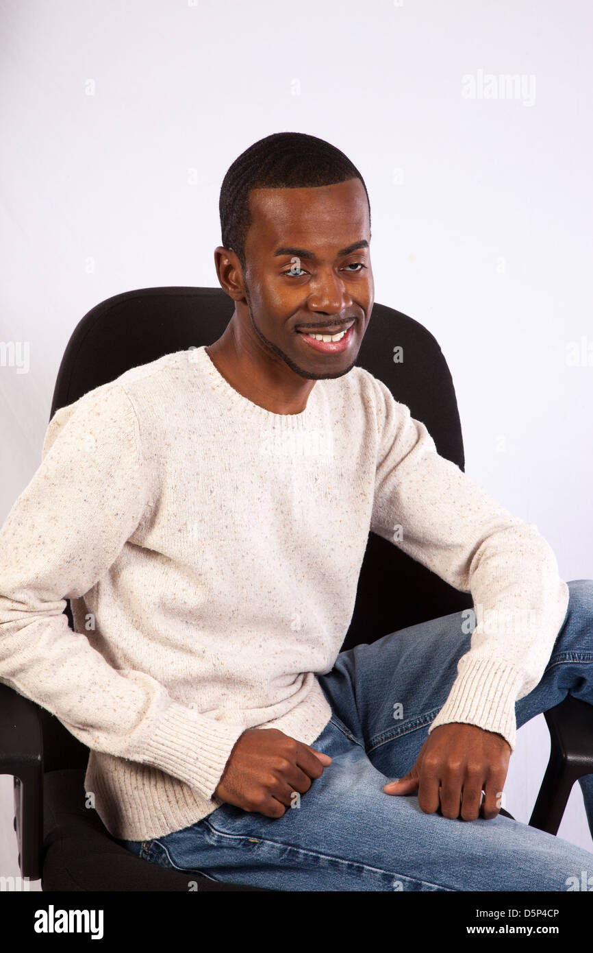Handsome black man  in pullover shirt and blue jeans, sitting casually in a business chair, smiling Stock Photo