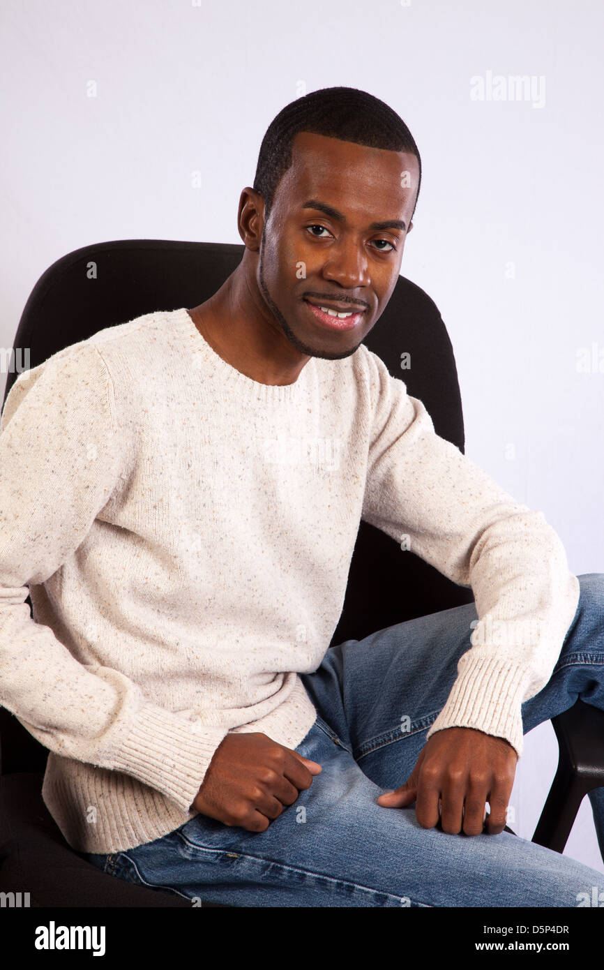 Handsome black man  in pullover shirt and blue jeans, sitting casually in a business chair, smiling at the camera Stock Photo