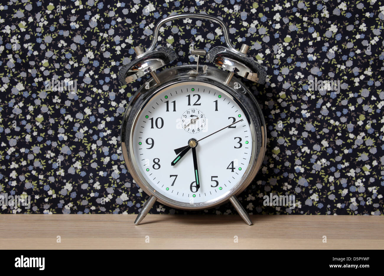 Old style alarm clock, on a dresser in front of floral wallpaper. Time set at 7.30 Stock Photo