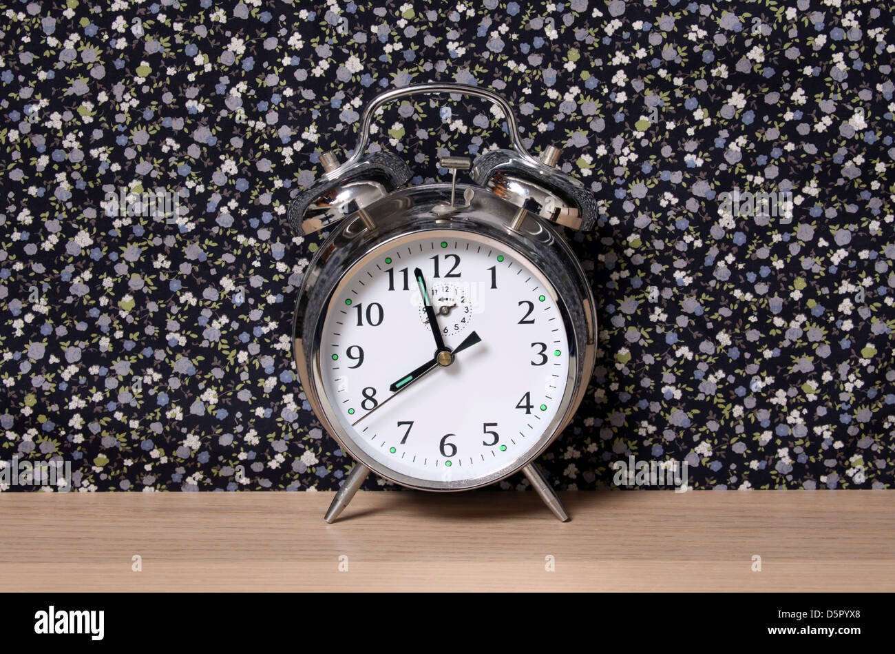 Old style alarm clock, on a dresser in front of floral wallpaper. Time set at just before 8. Stock Photo
