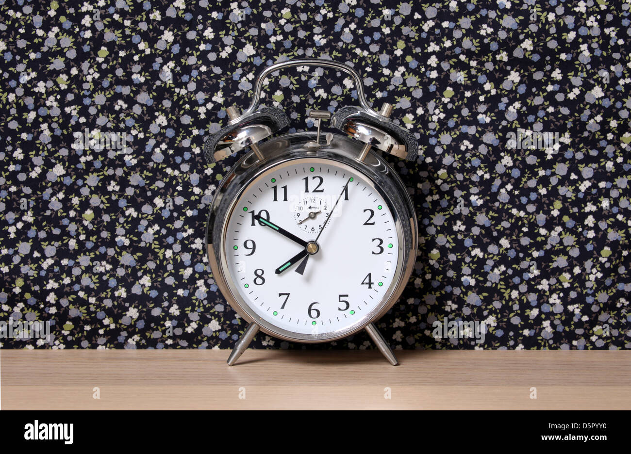 Old style alarm clock, on a dresser in front of floral wallpaper. Time set at 7.50. Stock Photo