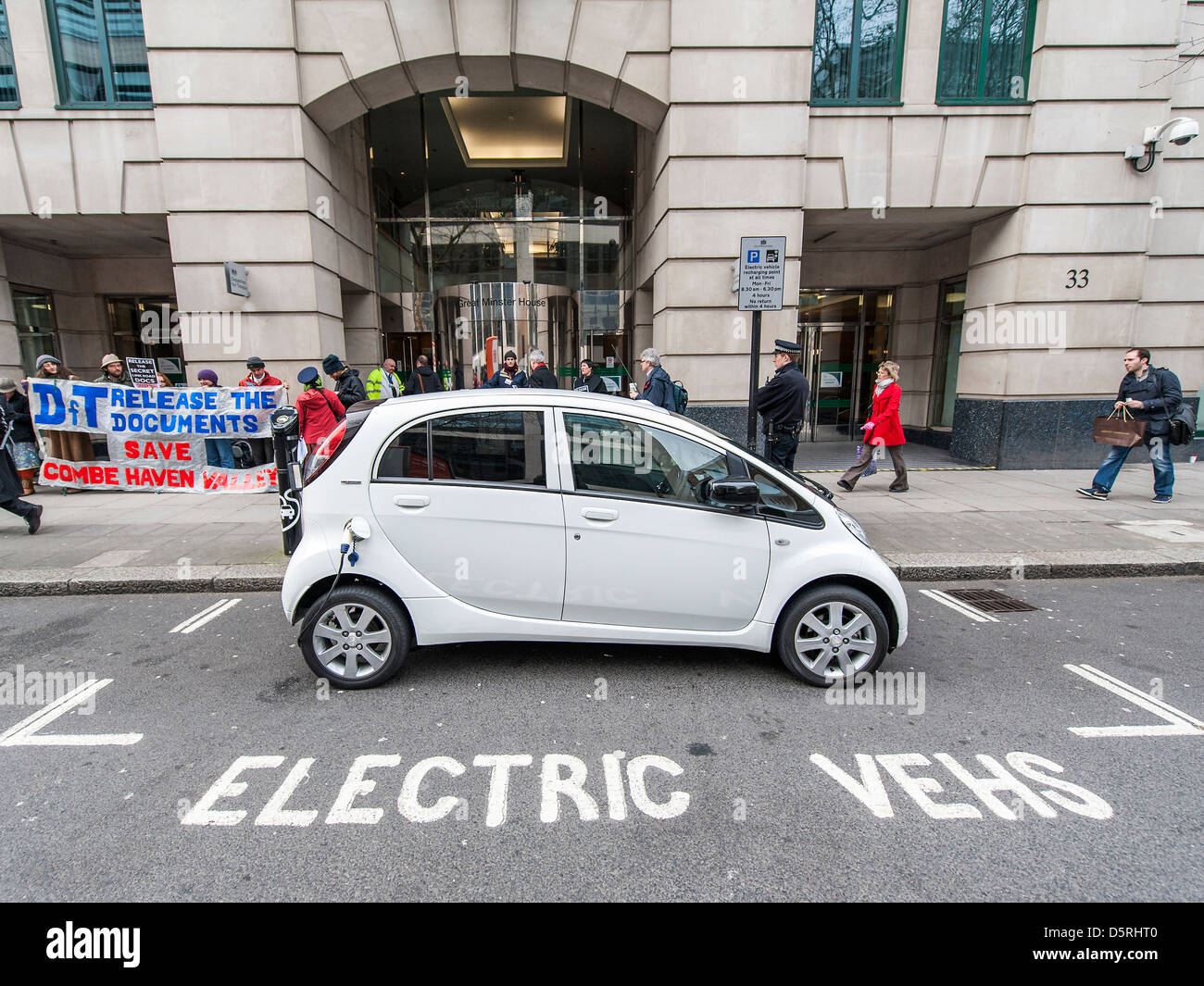 An electric car is charged outside the Department for Transport.  An anti road protest goes on in the background. Stock Photo