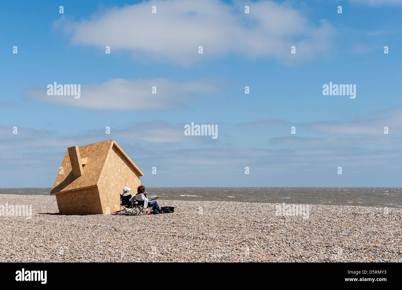 Two people sitting next to a wooden modern art sculpture of the House in the Clouds on Thorpeness beach in Suffolk, England, UK Stock Photo