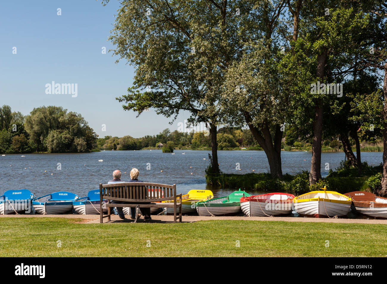 A couple sitting on a wooden bench overlooking Thorpeness Meare - a traditional boating lake in Suffolk, England, UK Stock Photo
