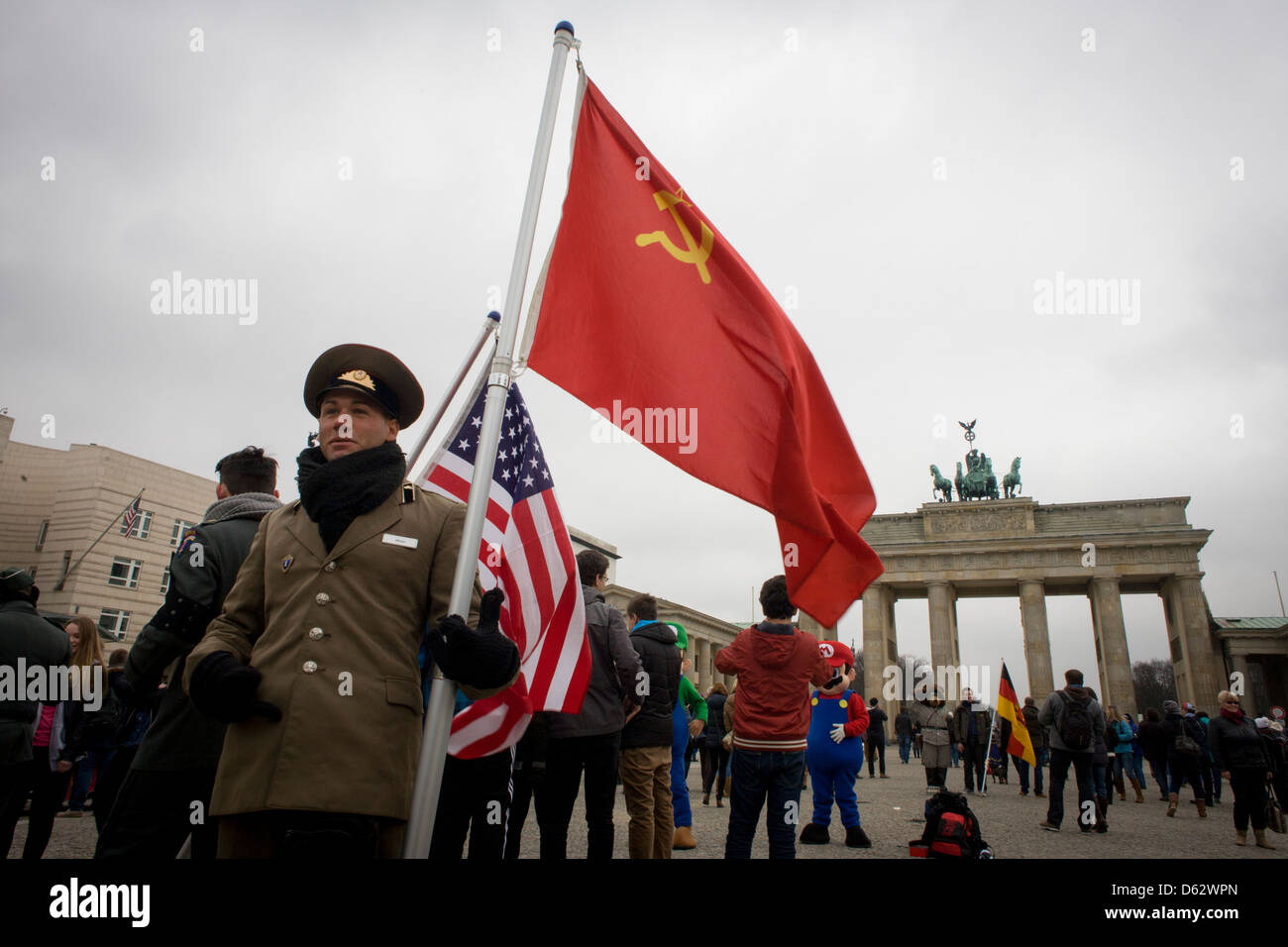 Actors in US and Soviet army uniforms hold flags to recount German history during the second world war and later, the cold war - beneath the Brandenburg Gate in Unter den Linden in central Berlin, Germany. The site is near the former border between Communist East and West Berlin during the Cold War. Here also, Berlin was separated by the occupying sectors of US, British, French and Soviet forces after WW2. (More in Description) .. Stock Photo