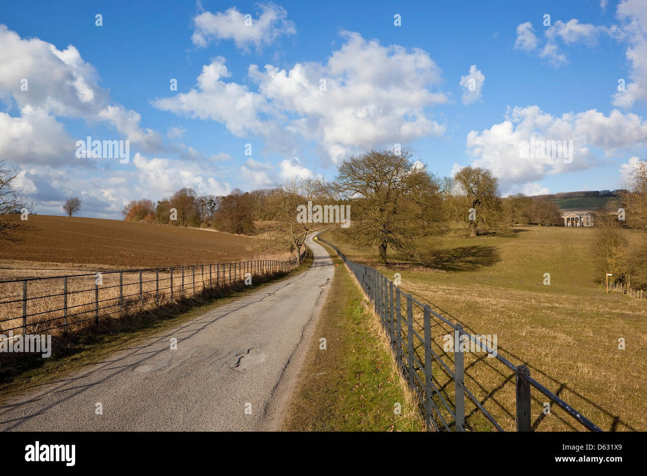 Yorkshire wolds landscape with a road through scenic parkland with iron railings and trees under a blue sky with clouds. Stock Photo