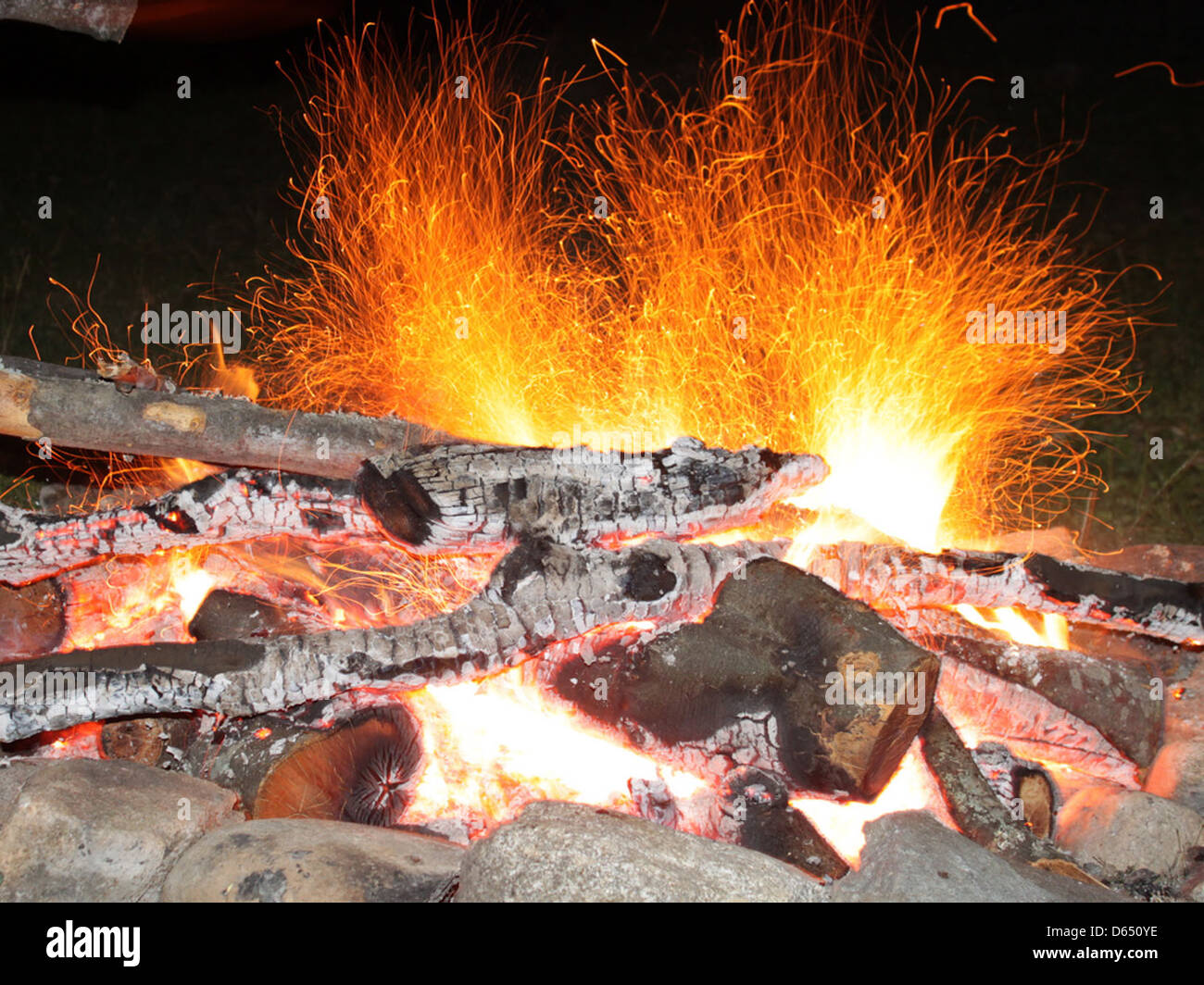 The image shows a campfire in the dark of night with sparks rising into ...