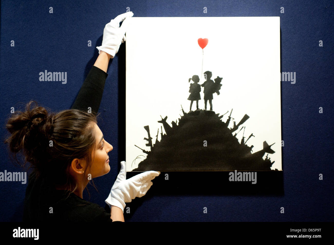 London, UK. 12th April 2013. a sales assistant holds a canvas entitled 'Kids on Guns' by Banksy during the 'Urban Art' auction preview at Bonhams. “Kids on Guns” is an example of the artist in more political mood, this canvas, which rarely appears at auction, being offered at £50,000-70,000. Like many of Banksy’s most famous creations, at first glance the painting may seem rather sweet, but on closer inspections its vision is much more sinister. Credit: Piero Cruciatti / Alamy Live News Stock Photo
