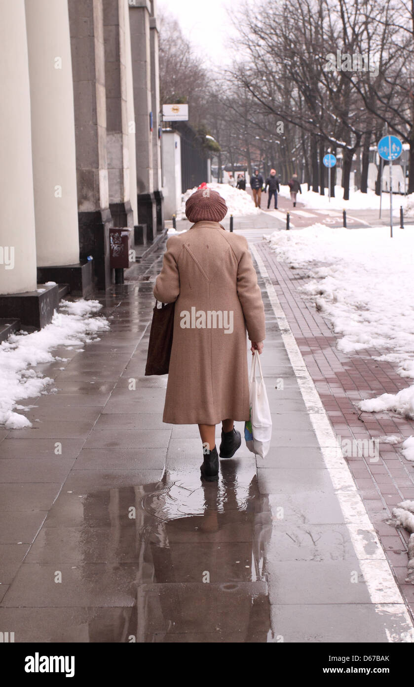 Old elderly woman carrying bags walking on a wet pavement with snow ...