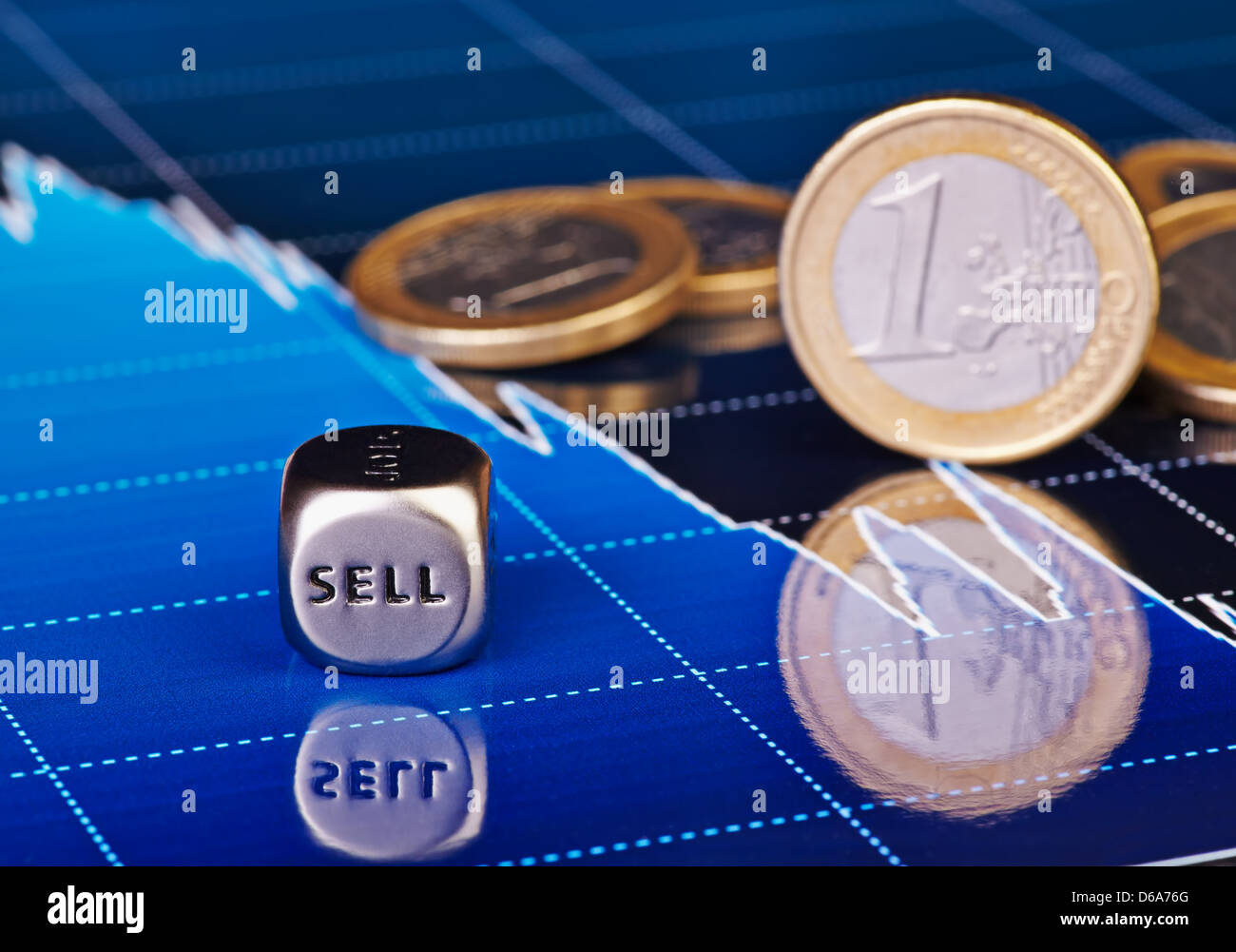 Close-up dices cube with the word SELL, one-euro coins and downtrend financial chart as background. Selective focus. Stock Photo