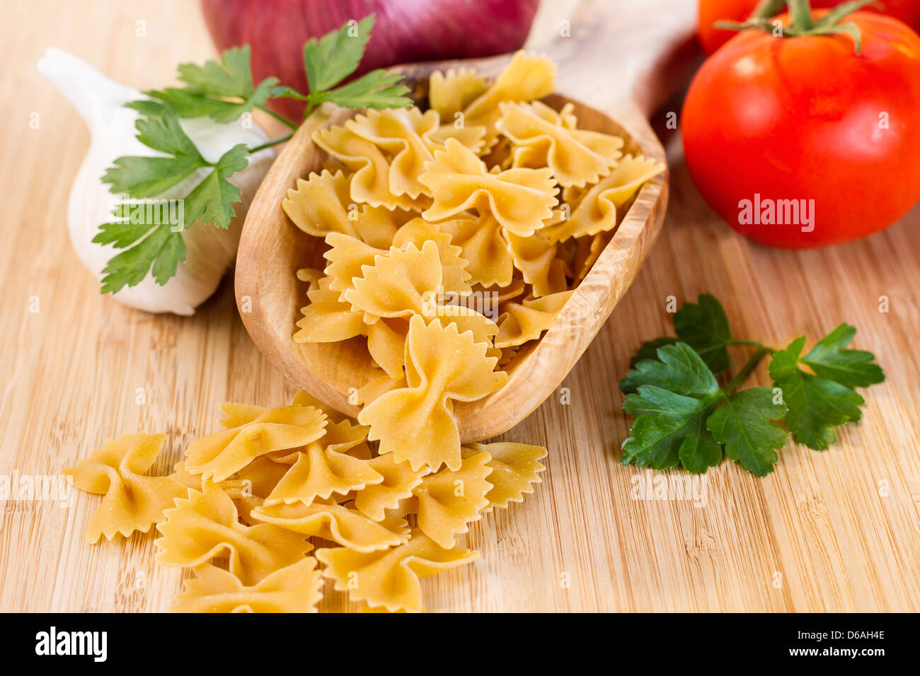 Closeup Horizontal photo of raw pasta in wooden spoon surround by onion, parsley, garlic and tomatoes on natural bamboo board Stock Photo