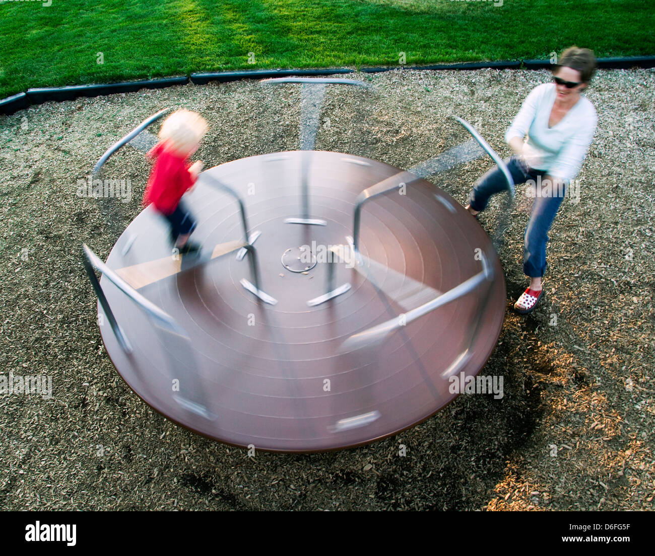 Young mother spinning her blonde haired boy on a merry-go-round on a playground. Stock Photo
