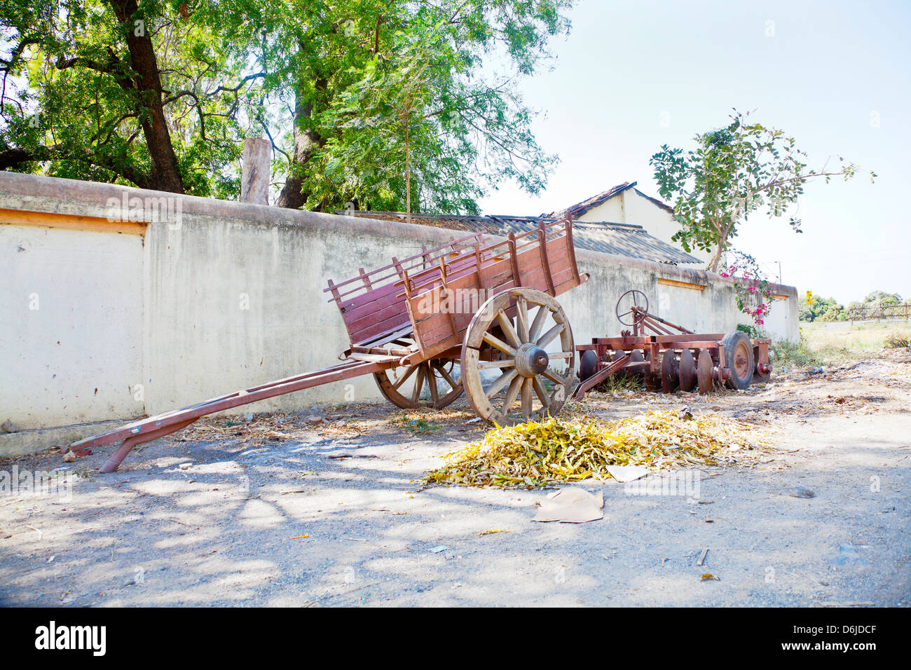 Outskirts of an Indian hinterlands village of a wall lined with farm machinery and parked bullock cart under the shade of trees Stock Photo