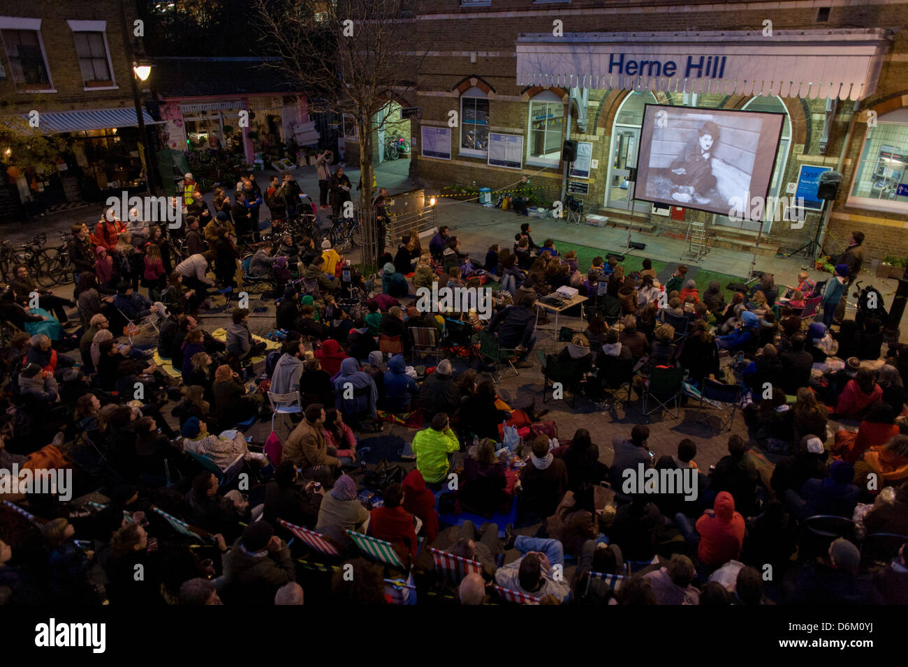 London, UK. 19th April, 2013.  Accompanied by a pianist, south Londoners sit outside their rail station on whose wall is a screening of the silent film Easy Street starring a former local boy, Charlie Chaplin, kicking off a series of the Free Film Festival in Herne Hill in the London borough of Lambeth. There is no official record of his birth although Chaplin believed he was born at East Street in nearby Walworth. Credit Richard Baker / Alamy Live News Stock Photo