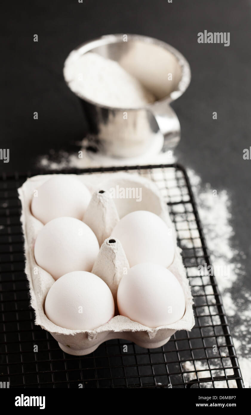 Chicken eggs in white carton on mesh tray with flour in metal measuring cup on dark background Stock Photo