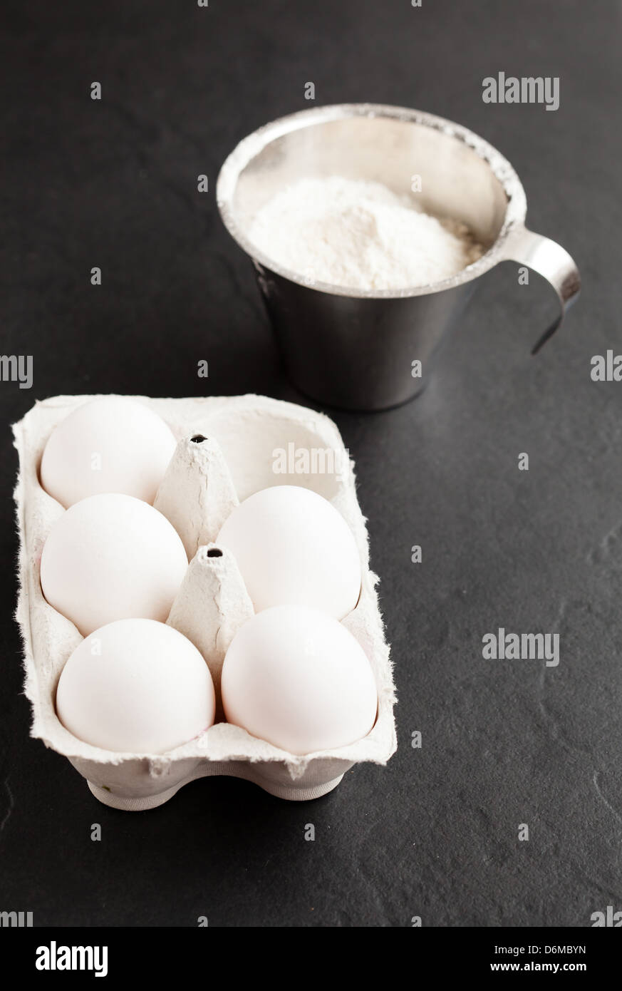 Chicken eggs in white carton with flour in metal measuring cup on dark background Stock Photo
