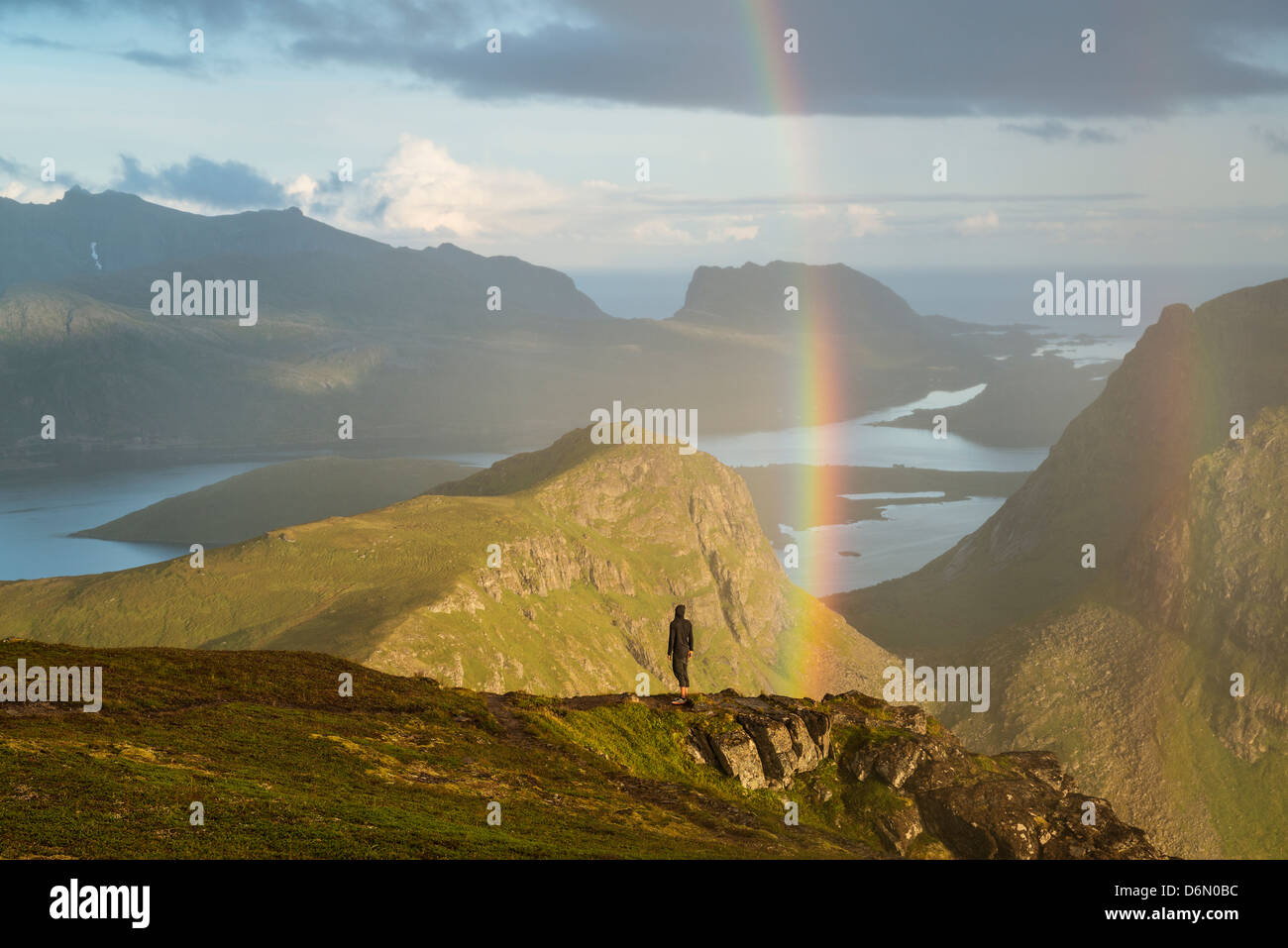 Person stands under rainbow near summit of Ryten, Lofoten Islands, Norway Stock Photo