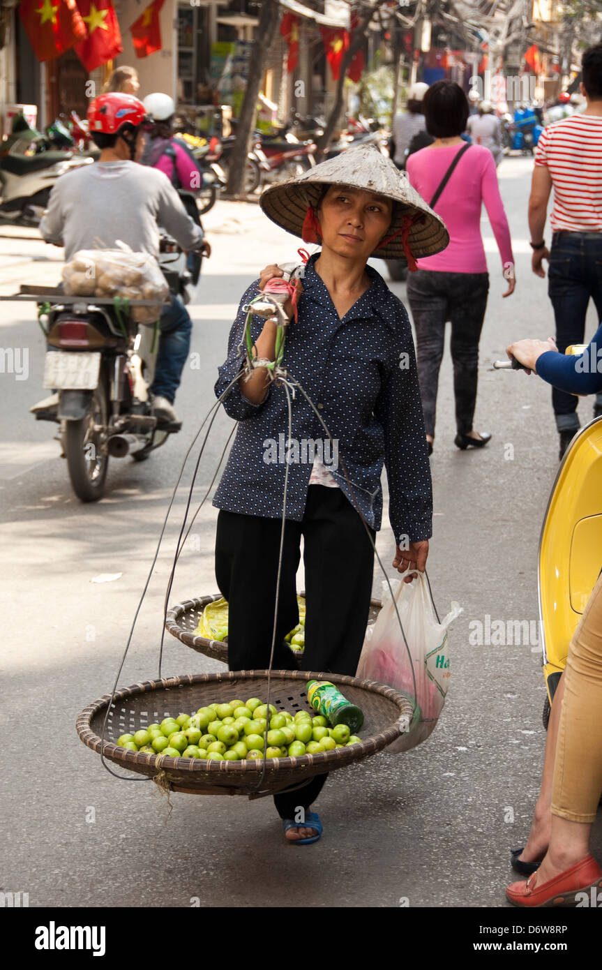 Vertical portrait of a Vietnamese lady carrying baskets of apples on a bamboo shoulder brace through the Old Quarter in Hanoi. Stock Photo